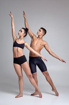 A pair of graceful modern ballet dancers in black suits are posing over a gray studio background. Handsome man in black shorts and beautiful woman in a black swimwear are dancing together. Ballet and contemporary choreography concept. Art photo.