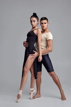 Pair of a modern ballet dancers posing over a gray studio background and looking at the camera. Handsome man in black shorts with beige t-shirt and beautiful woman in a black dress and white pointe shoes are dancing together. Ballet and contemporary choreography concept. Art photo.