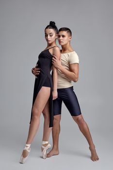 Couple of a young modern ballet dancers posing over a gray studio background and looking at the camera. Good-looking man in black shorts with beige t-shirt and beautiful woman in a black dress and white pointe shoes are dancing together. Ballet and contemporary choreography concept. Art photo.