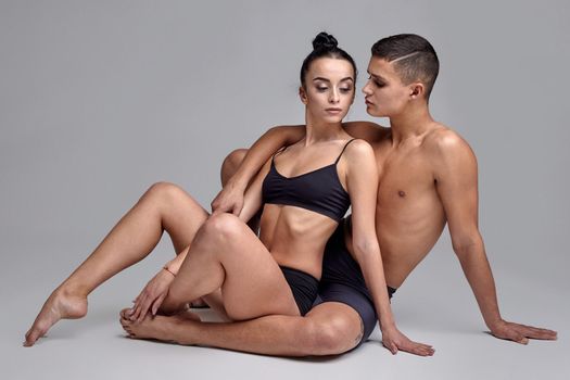A pair of an athletic ballet dancers in black suits are posing over a gray studio background. Handsome male in black shorts is embracing a sexy woman in a black swimwear. Ballet and contemporary choreography concept. Art photo.