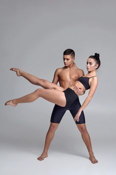 Two young modern ballet dancers in black suits are posing over a gray studio background. Handsome man in black shorts is holding a beautiful woman in a black swimwear. Ballet and contemporary choreography concept. Art photo.