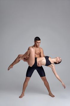 Two young ballet dancers in black suits are posing over a gray studio background. Attractive male in black shorts is holding a beautiful female in a black swimwear. Ballet and contemporary choreography concept. Art photo.