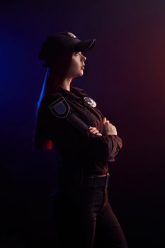 Pretty redheaded woman police officer in a uniform and a cap is posing sideways with crossed hands and looking away, against a black background with red and blue backlighting.