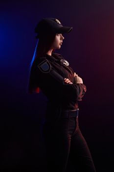 Pretty redheaded female police officer in a uniform and a cap is posing sideways with crossed hands and looking away, against a black background with red and blue backlighting.