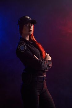 Attractiveginger girl police officer in a uniform and a cap is posing sideways with crossed hands and looking away, against a black background with red and blue backlighting.
