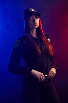 Alluring ginger girl police officer in a uniform and a cap, with bright make-up, is holding her belt and looking at the camera against a black background with red and blue backlighting. Defender of citizens is ready to enforce a law and stop a crime.