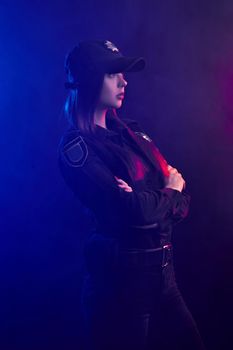Alluring redheaded woman police officer in a uniform and a cap, with bright make-up, is standing sideways and looking away against a black background with red and blue backlighting.