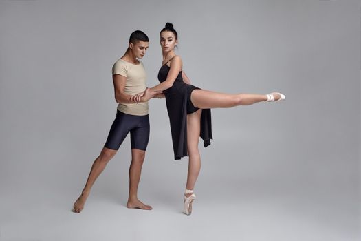 Full length photo of a professional ballet dancers posing over a gray studio background. Handsome man in black shorts with beige t-shirt and beautiful woman in a black dress and white pointe shoes are dancing together. Ballet and contemporary choreography concept. Art photo.