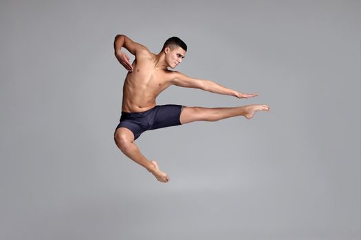 Studio shot of a stately male ballet dancer, dressed in a black shorts. He is jumping on a gray background in studio. Bare legs and torso. Ballet and contemporary choreography concept. Art photo.