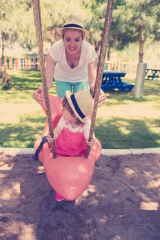 young mother and her little daughter smiling together while swinging at the playground in the park