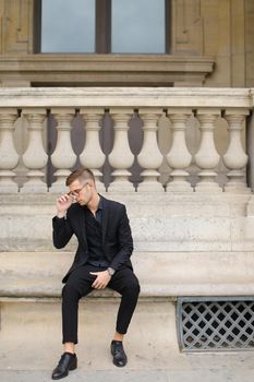 Young man sitting on sidewalk and leaning on concrete railing of building, wearing black suit and glasses. Concept of walkig in city, urban photo session and male person model.