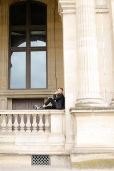 Young caucasian european boy sitting on balcony concrete banister in Paris. Concept of architecture and male fashion model. Guy wearing black suit and walking in city.