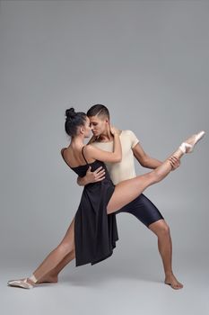 Full length studio shot of a modern ballet dancers posing over a gray background. Strong male in black shorts with beige t-shirt is holding a charming girl in a black dress and white pointe shoes in his strong hands. Ballet and contemporary choreography concept. Art photo.