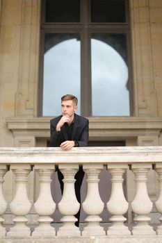 Young european handsome boy leaning on balcony concrete railing in Paris. Concept of male fashon moden and architecture. Guy wearing black suit.