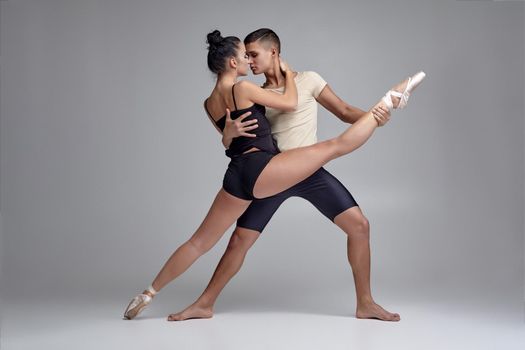 Studio portrait of a creative ballet dancers posing over a gray studio background. Attractive man in black shorts with beige t-shirt is holding a lovely girl in a black dress and white pointe shoes in his strong hands. Ballet and contemporary choreography concept. Art photo.