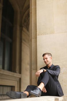 Young caucasian boy sitting on balcony concrete banister in Paris. Concept of architecture and male fashion model. Guy wearing black suit and walking in city.