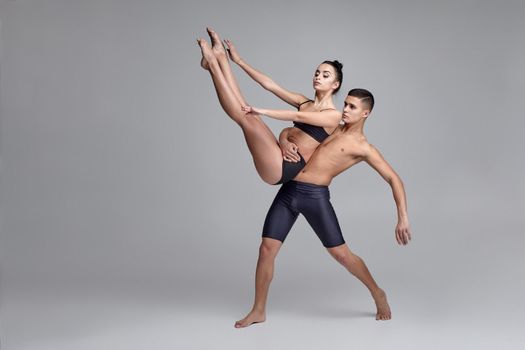 A couple of a professional ballet dancers in black suits are posing over a gray studio background. Good-looking man in black shorts is holding in his hands a beautiful woman in a black swimwear. Ballet and contemporary choreography concept. Art photo.