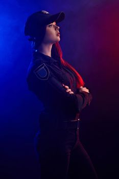 Alluring redheaded lady police officer in a uniform and a cap is posing sideways with crossed hands and looking away, against a black background with red and blue backlighting. Defender of citizens is ready to enforce a law and stop a crime.