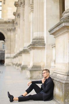 Young caucasian man sitting on sidewalk ground and wearing black suit, leaning on concrete banister. Concept of walking in cty and male fashionable model.