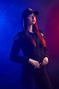 Attractive ginger girl police officer in a uniform and a cap, with bright make-up, is standing sideways, holding her belt and looking away against a black background with red and blue backlighting.