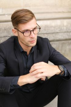 Portrait of man wearing black suit and glasses, sitting on sidewalk ground. Concept of male beauty and fashionable model.