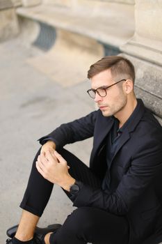 Young fashionable man sitting on sidewalk ground and wearing black suit, leaning on concrete banister. Concept of walking in cty and male stylish model.