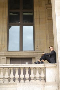 Young caucasian handsome boy sitting on balcony concrete banister in Paris. Concept of architecture and male fashion model. Guy wearing black suit and walking in city.