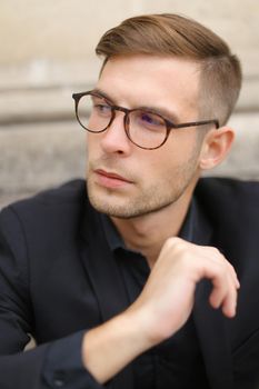 Closeup portrait of man wearing black suit and glasses, sitting on sidewalk ground. Concept of male beauty and fashionable model.