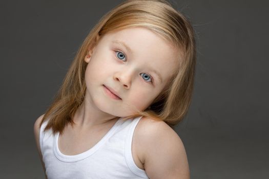 Calm cute little girl wearing a white undershirt looking at the camera while standing on grey background. Childhood concept