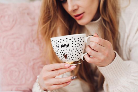 Close-up stock photo of a young fair-haired woman holding a cute coffee cup with golden design and text be mine . Morning coffee in a beautiful coffee cup.