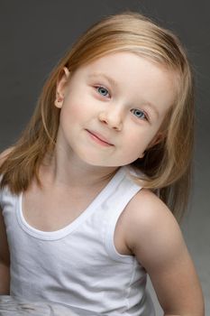 Cute fair haired girl in a white undershirt standing on dark grey background and smiling. Childhood concept