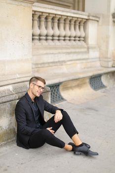 Young handsome man sitting on sidewalk ground and wearing black suit, leaning on concrete banister. Concept of walking in cty and male fashionable model.