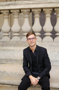 Young handsome man sitting on sidewalk and leaning on concrete railing of building, wearing black suit and glasses. Concept of walkig in city, urban photo session and male person model.