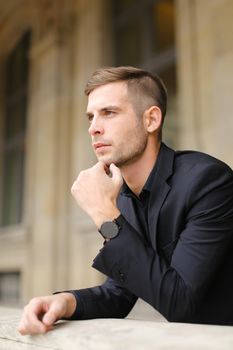 Closeup portrait of young businessman on balcony leaning on banister. Concept of male model and fashion. Young man wearing black suit.