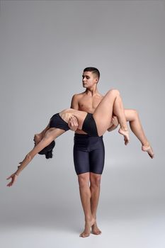Two young professional ballet dancers in black suits are posing over a gray studio background. Handsome male in black shorts is holding a charming female in a black swimwear. Ballet and contemporary choreography concept. Art photo.