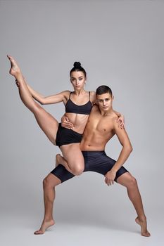 A pair of a strong ballet dancers in black suits are posing over a gray studio background. Athletic male in black shorts is holding in his hands a pretty girl in a black swimwear. Ballet and contemporary choreography concept. Art photo.