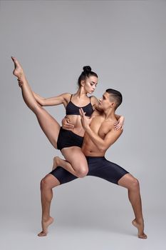 A pair of a strong ballet dancers in black suits are posing over a gray studio background. Athletic man in black shorts is holding in his hands a lovely girl in black swimwear and they are looking at each other. Ballet and contemporary choreography concept. Art photo.