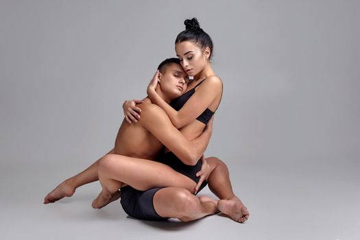 A pair of loving ballet dancers in black suits are posing over a gray studio background. Attractive man in black shorts and charming woman in a black swimwear are embracing each other. Contemporary choreography concept. Art photo.