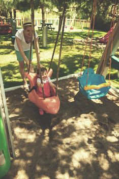 young mother and her little daughter smiling together while swinging at the playground in the park