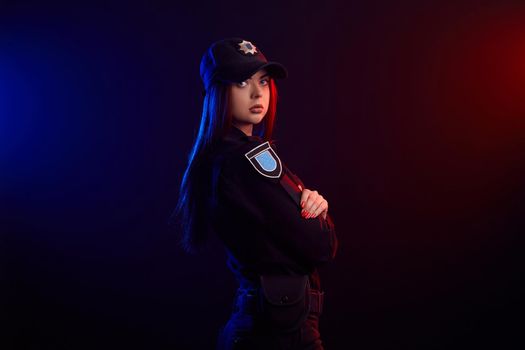 Serious redheaded female police officer in a uniform and a cap, with bright make-up, is posing sideways and looking at the camera, against a black background with red and blue backlighting. Defender of citizens is ready to enforce a law and stop a crime.