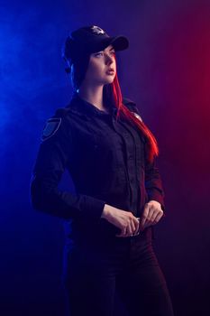 Serious redheaded woman police officer in a uniform and a cap, with bright make-up, is holding her belt and looking away against a black background with red and blue backlighting.