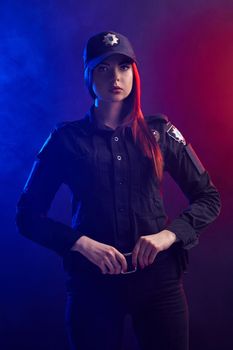 Serious redheaded female police officer in a uniform and a cap, with bright make-up, is holding her belt and looking at the camera against a black background with red and blue backlighting.