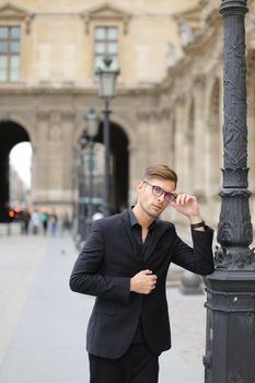 Young european man walking in Paris and leaning on lantern, weaaring black suit. Concept of walking in city and male fashion.