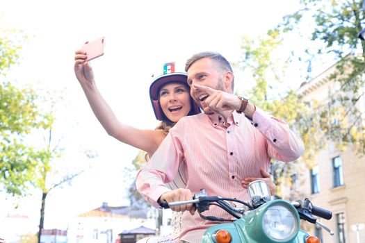 Happy young couple taking selfie on smartphone while sitting on scooter outdoors