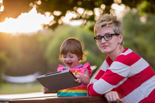 Happy mother and her little daughter enjoying free time using tablet computer while relaxing  on holiday home garden during sunny day