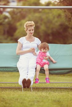 A beautiful young mother and her happy little daughter enjoying free time while swinging on bench at backyard