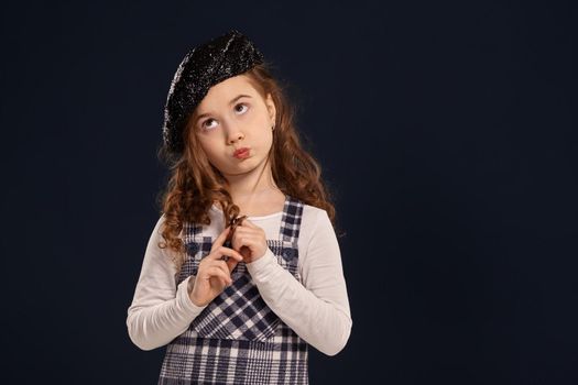 Nice little girl with a beautiful curly hair wearing checkered dress, white blouse and a black sparkling beret is looking thoughtful. Stylish brunette kid is posing in studio on a black background. Children's fashion.