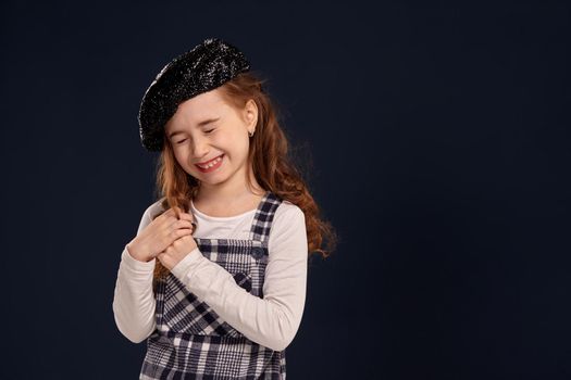 Emotional little girl with a beautiful curly hair wearing checkered dress, white blouse and a black sparkling beret has closed her eyes and laughing. Stylish brunette kid is posing in studio on a black background. Children's fashion.