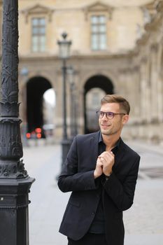 Young man in glasses walking in Paris and wearing black suit, standing near lantern, France. Concept of walking in city and male fashion.