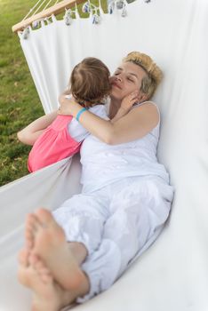 Happy mother and a little daughter enjoying free time hugging and relaxing in a hammock during a sunny day on holiday home garden